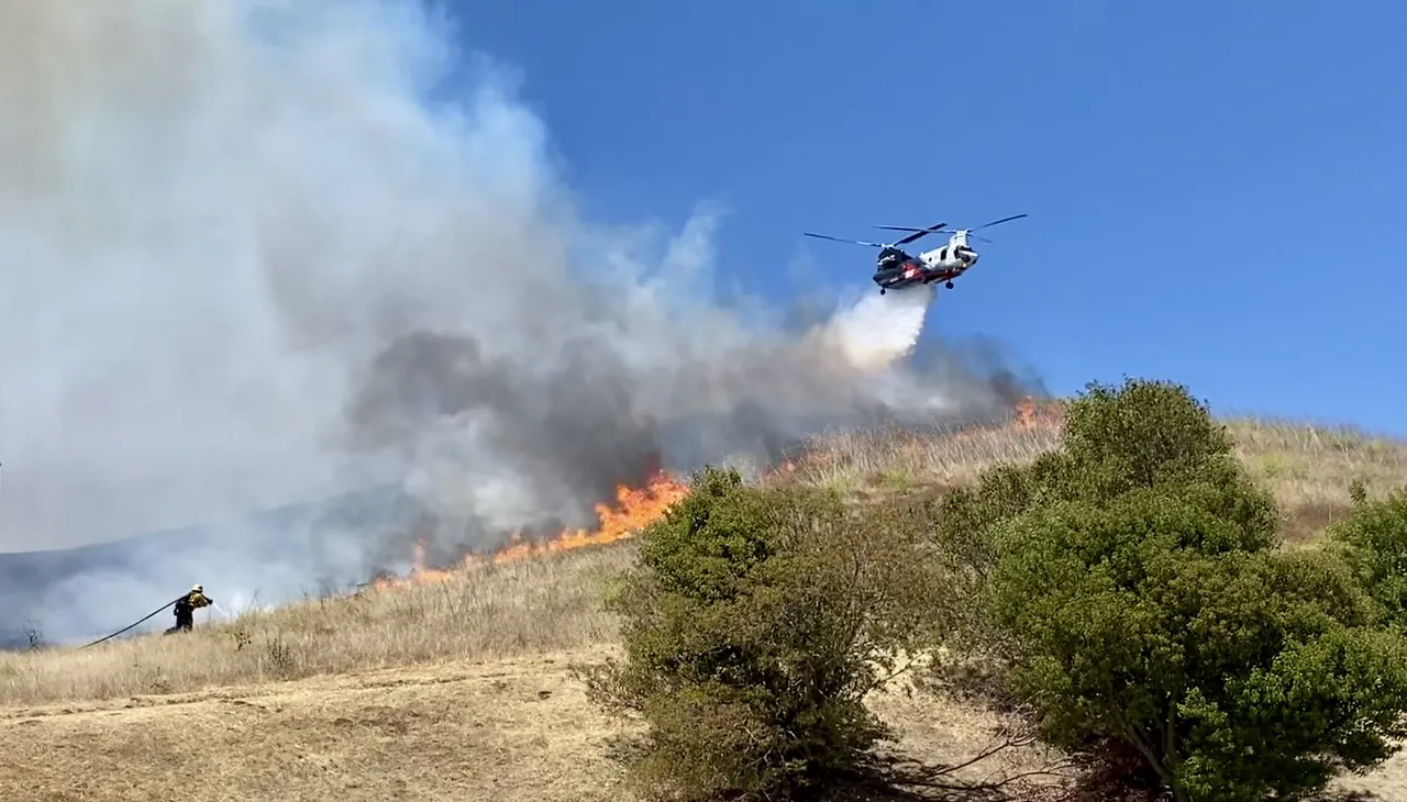 A helicopter drops water on the Sandy Fire burning across a dry chaparral hillside near Simi Valley, May 2026
