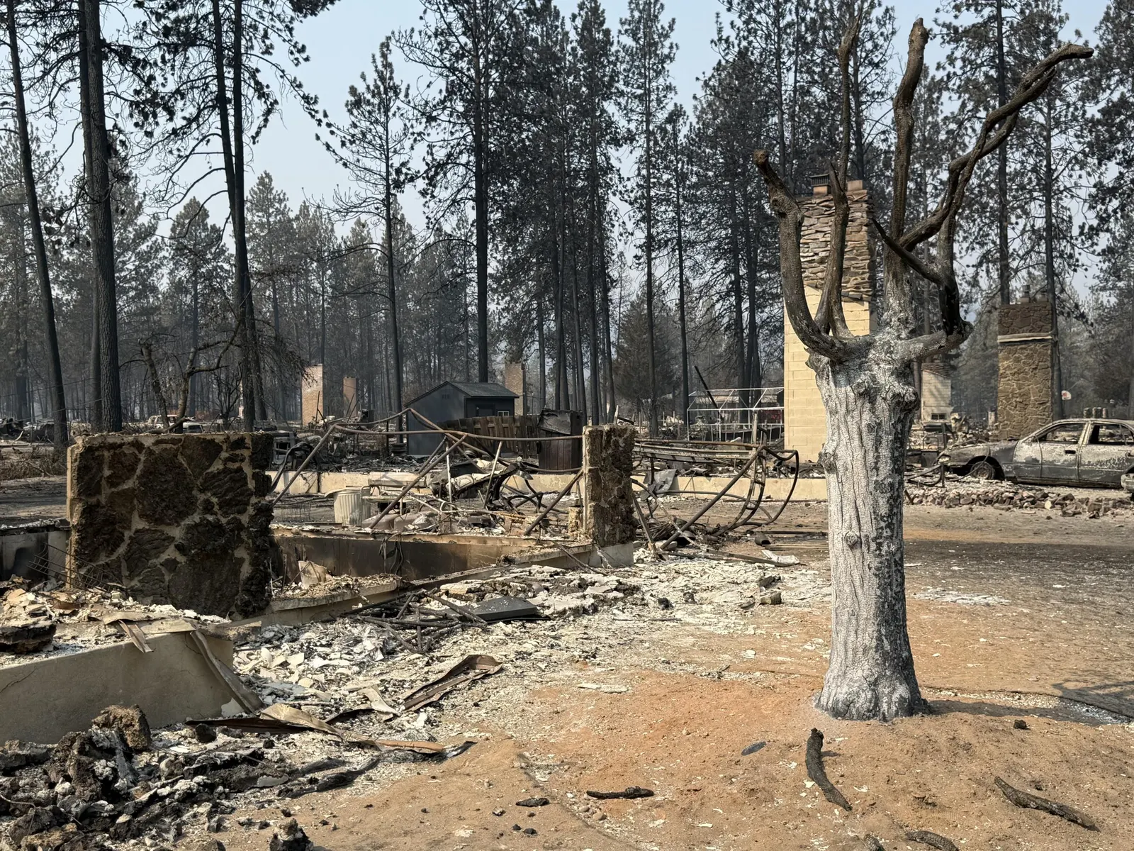 A burned neighborhood in the Old Trails Fire scar near Spokane, Washington, August 2026 — stone chimneys, a destroyed car and scorched ponderosa pines around leveled homes